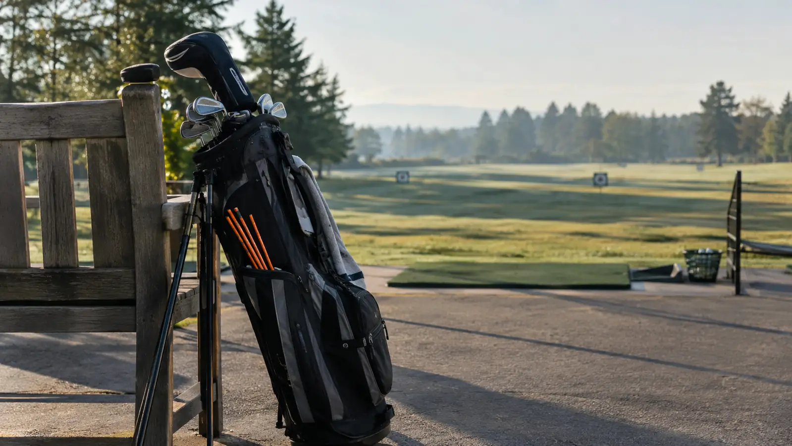 A golf bag leaning against a bench at a driving range in the morning, with a set of collapsible alignment sticks tucked into the side pocket.