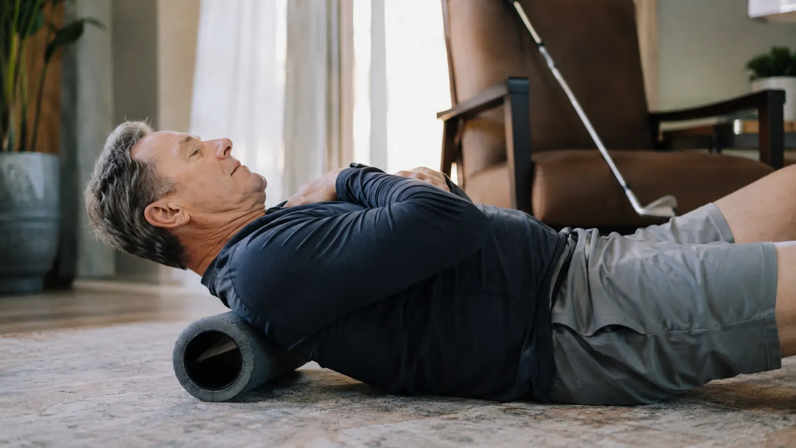 A man in his sixties kneeling on a living room rug with a foam roller under his upper back, a golf club leaning against a chair nearby